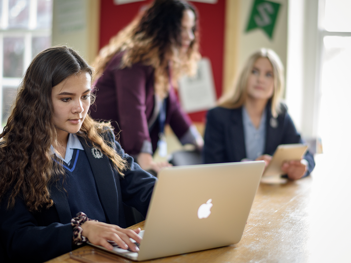 Students using mac laptops to work in a classroom, overseen by a teacher